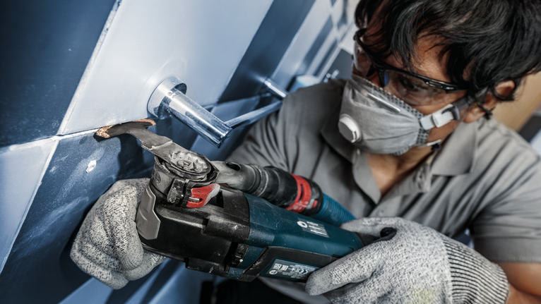 Una persona che indossa un equipaggiamento di sicurezza utilizza un elettroutensile per tagliare le piastrelle attorno a un tubo.