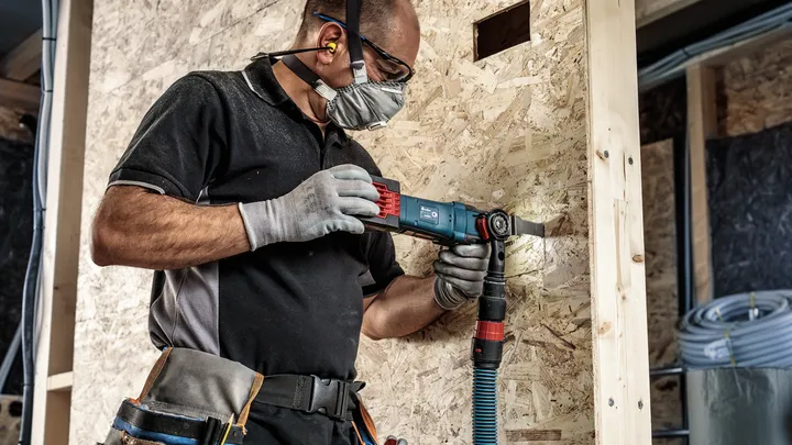 Una persona che indossa dispositivi di sicurezza taglia un pannello di legno con un elettroutensile.