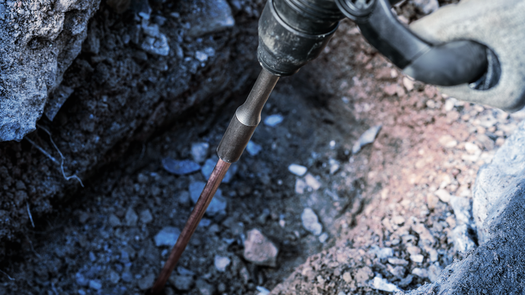 Una persona che indossa un equipaggiamento di sicurezza utilizza un elettroutensile per rompere il terreno vicino alle rocce.