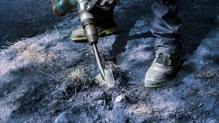Una persona che indossa un equipaggiamento di sicurezza utilizza un elettroutensile con uno scalpello per rompere il terreno.