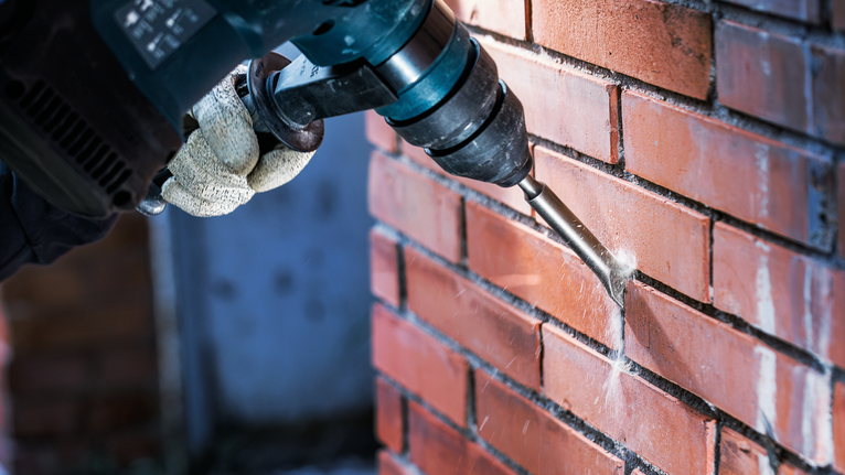 Una persona che indossa un equipaggiamento di sicurezza fora un muro di mattoni con un utensile elettrico.