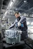 Una persona che indossa dispositivi di sicurezza utilizza un trapano a batteria per assemblare macchinari in una fabbrica.