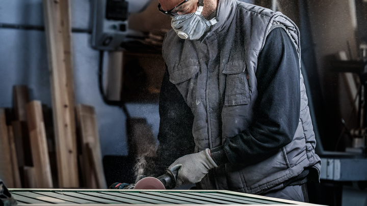 Una persona che indossa un equipaggiamento di sicurezza leviga una superficie di legno con un utensile elettrico.