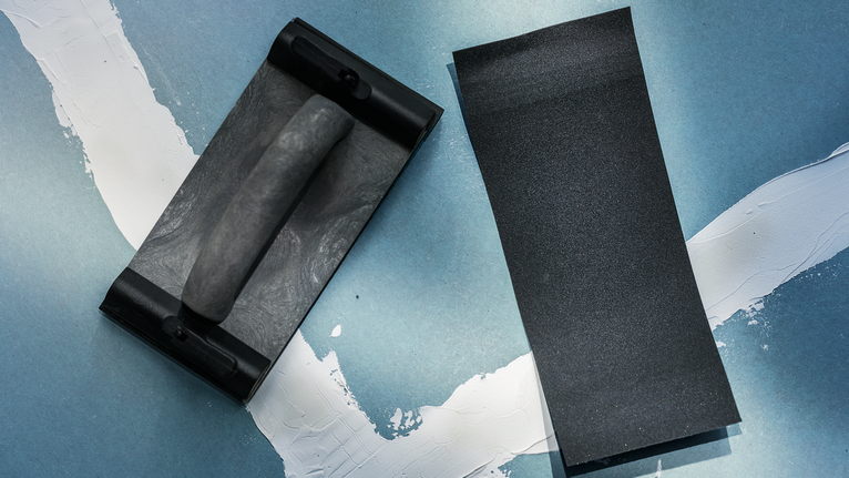 Blocco abrasivo portatile e un foglio di carta vetrata su una superficie dipinta di blu.