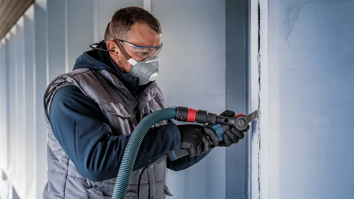 Una persona che indossa un equipaggiamento di sicurezza esegue una scanalatura verticale in un muro utilizzando un utensile elettrico.