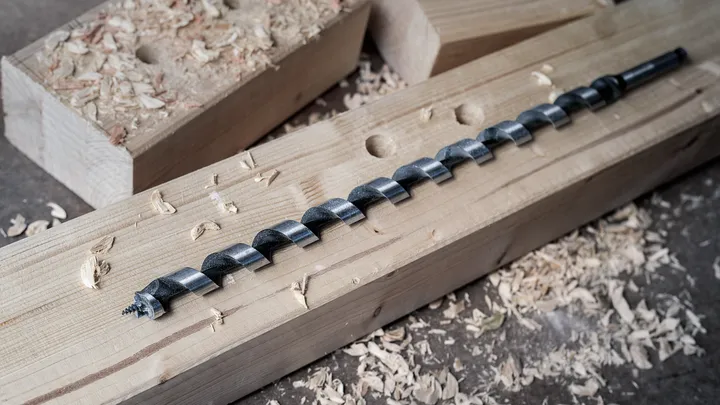 Punta lunga da trapano su una trave di legno circondata da trucioli di legno.