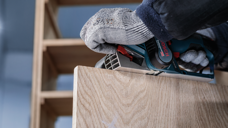 Una persona che indossa l'equipaggiamento di sicurezza utilizza una pialla elettrica su una tavola di legno.