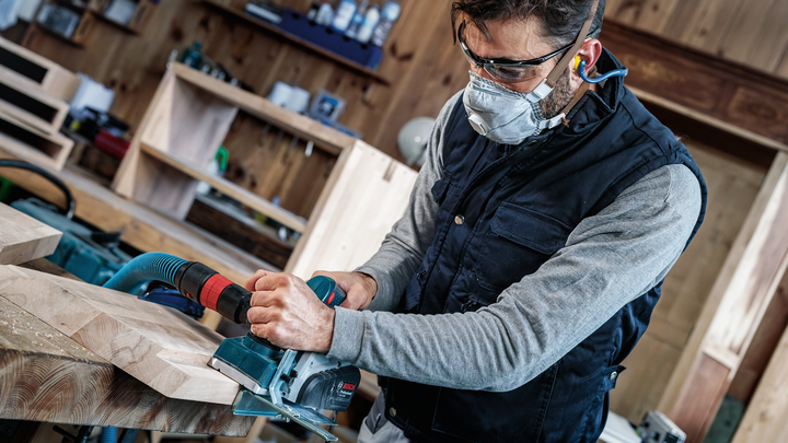 Una persona che indossa dispositivi di sicurezza utilizza una pialla elettrica per levigare le assi di legno.