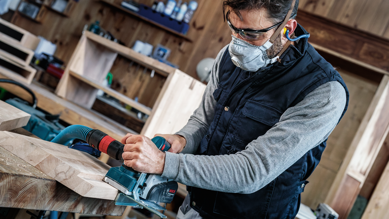 Una persona che indossa dispositivi di sicurezza utilizza una pialla elettrica per levigare le assi di legno.