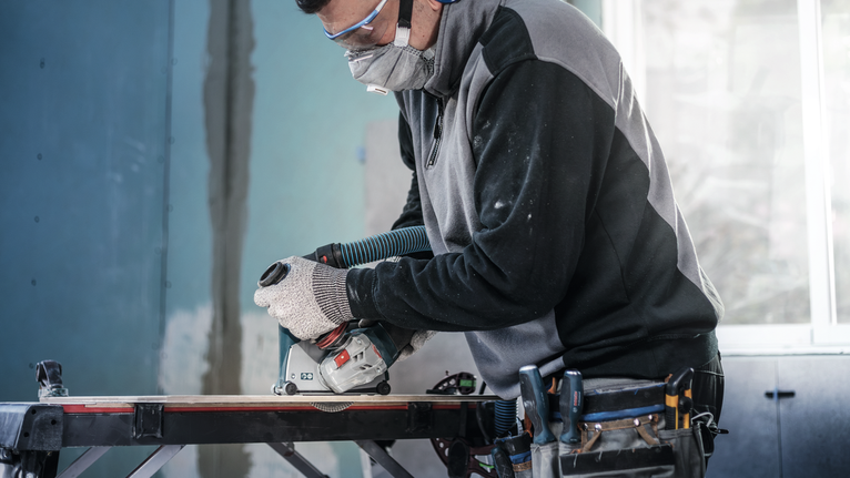 Una persona che indossa dispositivi di sicurezza utilizza una sega circolare per tagliare il legno su un banco da lavoro.