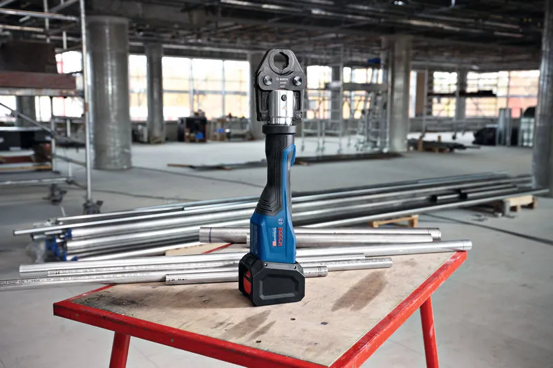 Utensile a pressa senza fili su un banco da lavoro circondato da tubi metallici in un cantiere edile.