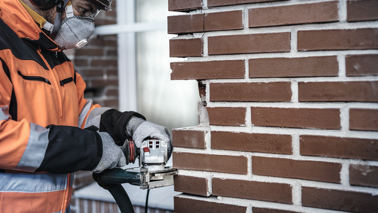 Una persona che indossa un equipaggiamento di sicurezza taglia una scanalatura in un muro di mattoni con un utensile elettrico.