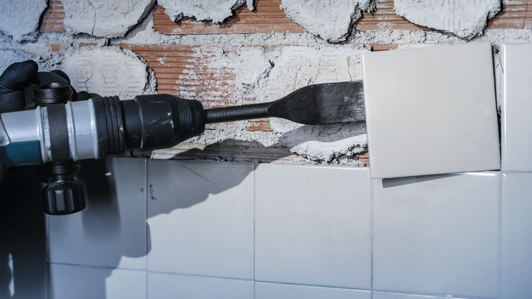 Una persona che indossa dispositivi di sicurezza utilizza un elettroutensile per rimuovere le piastrelle dal muro.