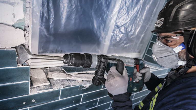 Un operaio che indossa dispositivi di sicurezza rimuove le piastrelle del muro utilizzando un martello demolitore.