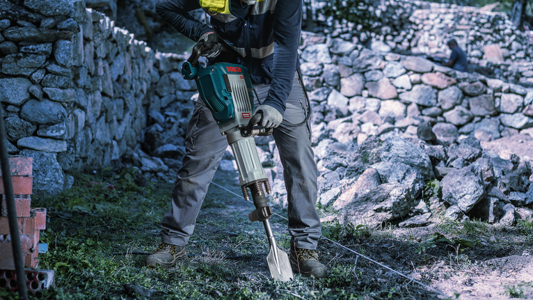 Una persona che indossa un equipaggiamento di sicurezza utilizza un martello demolitore per rompere il terreno vicino a un muro di pietra.