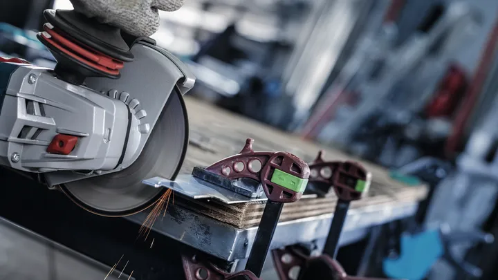Una persona che indossa un equipaggiamento di sicurezza taglia il metallo fissato a un banco da lavoro con una smerigliatrice angolare.
