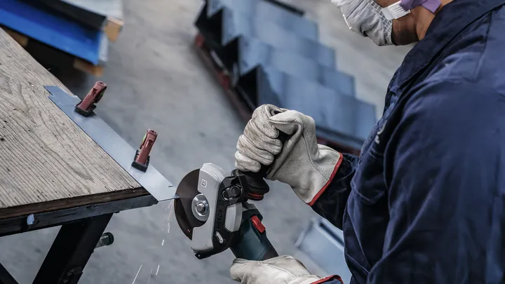 Una persona che indossa un equipaggiamento di sicurezza leviga una lamiera fissata a un banco da lavoro.