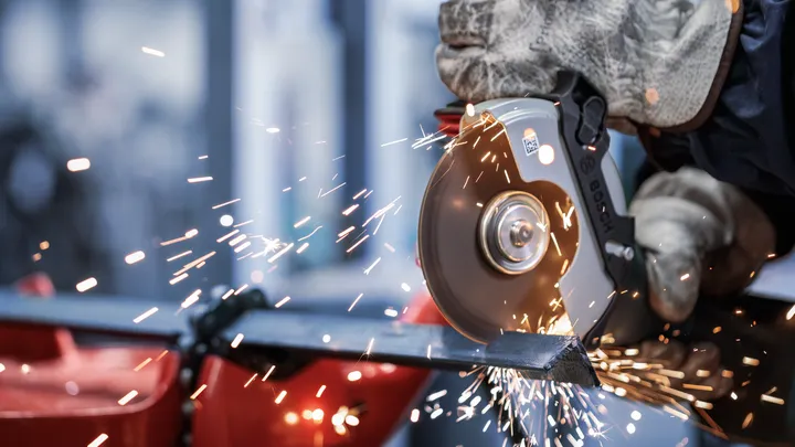 Una persona che indossa dispositivi di sicurezza utilizza una smerigliatrice angolare per tagliare il metallo, producendo scintille.