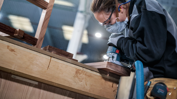 Una persona che indossa dispositivi di sicurezza utilizza un elettroutensile per levigare il bordo di una superficie in legno.