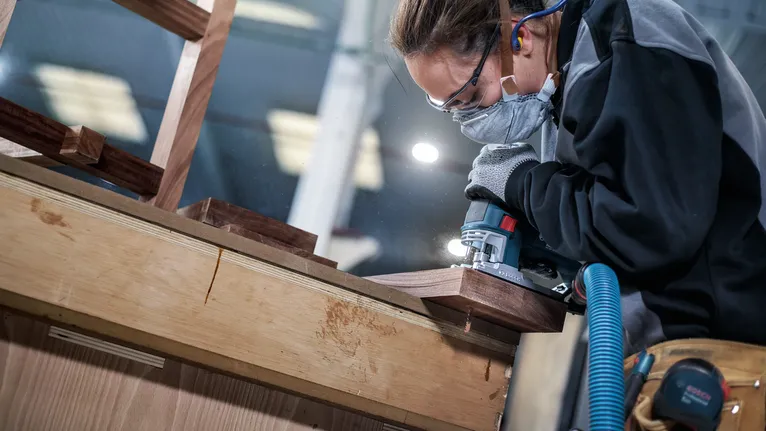 Una persona che indossa dispositivi di sicurezza utilizza un elettroutensile per levigare il bordo di una superficie in legno.