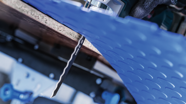 Una persona che indossa un equipaggiamento di sicurezza usa un seghetto alternativo per tagliare un foglio di plastica blu.