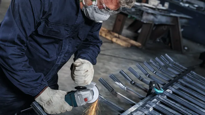 Una persona che indossa dispositivi di sicurezza rettifica barre di metallo con un utensile elettrico.