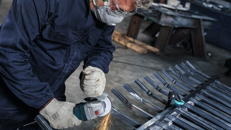 Una persona che indossa dispositivi di sicurezza rettifica barre di metallo con un utensile elettrico.