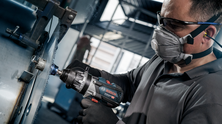 Una persona che indossa un equipaggiamento di sicurezza pratica un foro nel metallo utilizzando un utensile elettrico senza fili.