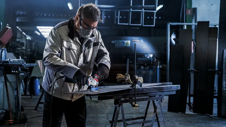 Una persona che indossa un equipaggiamento di sicurezza utilizza una smerigliatrice angolare su una superficie metallica.