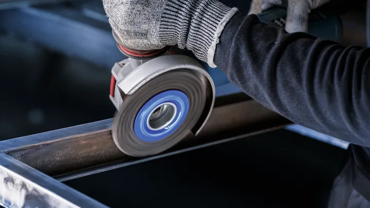 Una persona che indossa un equipaggiamento di sicurezza leviga una trave metallica con una smerigliatrice angolare.