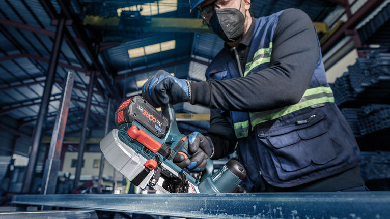 Una persona che indossa un equipaggiamento di sicurezza utilizza una sega a nastro senza fili per tagliare una trave di metallo.