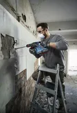 Una persona che indossa dispositivi di sicurezza rimuove le piastrelle del muro con un martello perforatore.