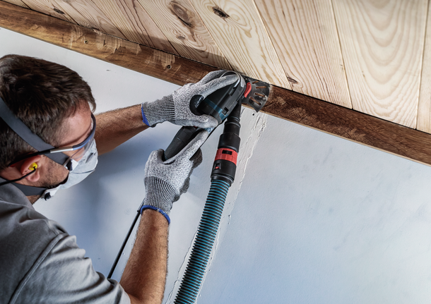 Una persona che indossa dispositivi di sicurezza leviga un angolo del soffitto in legno con un utensile elettrico.