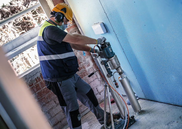 Una persona che indossa un equipaggiamento di sicurezza fora un muro utilizzando un elettroutensile montato.
