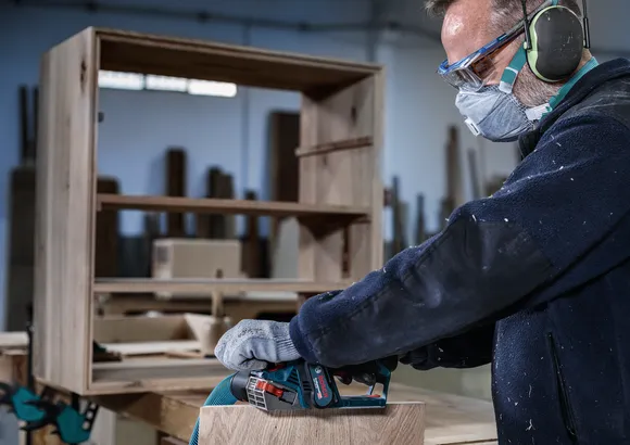 Una persona che indossa dispositivi di sicurezza leviga mobili in legno con un utensile elettrico.