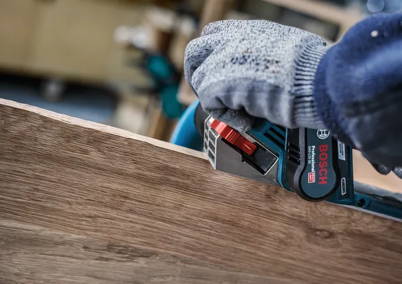 Una persona che indossa dispositivi di sicurezza leviga il bordo di una tavola di legno con un utensile elettrico.