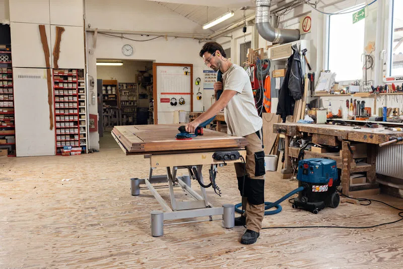 Una persona che indossa dispositivi di sicurezza leviga una superficie di legno con una levigatrice orbitale casuale a batteria.