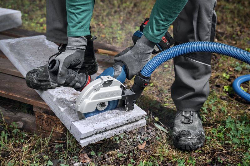 Una persona che indossa dispositivi di sicurezza utilizza una grande smerigliatrice angolare con aspirazione della polvere per tagliare il cemento.
