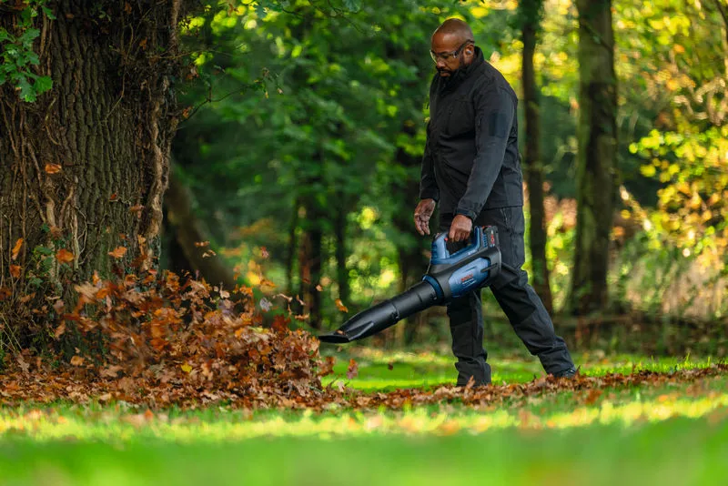 Una persona che indossa un equipaggiamento di sicurezza utilizza un soffiatore per rimuovere le foglie cadute vicino a un albero.