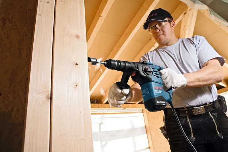 Una persona che indossa un equipaggiamento di sicurezza fora una trave di legno con un martello perforatore.