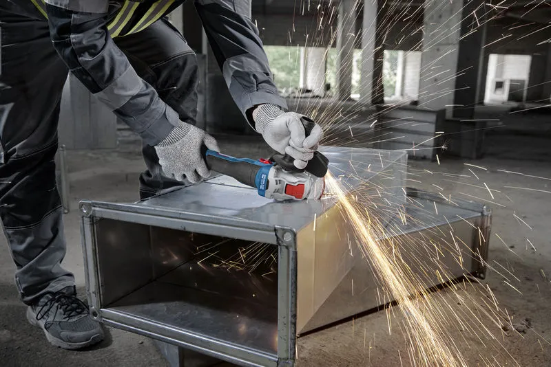 Una persona che indossa un equipaggiamento di sicurezza leviga un condotto metallico con una smerigliatrice angolare, producendo scintille.