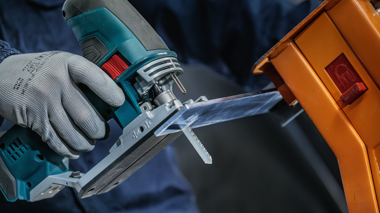 Una persona che indossa un equipaggiamento di sicurezza usa un seghetto alternativo per tagliare un foglio di plastica trasparente.