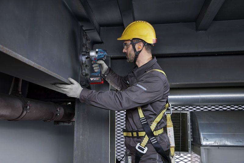 Una persona che indossa dispositivi di sicurezza stringe i bulloni su una trave di acciaio utilizzando una chiave a impulsi a batteria.