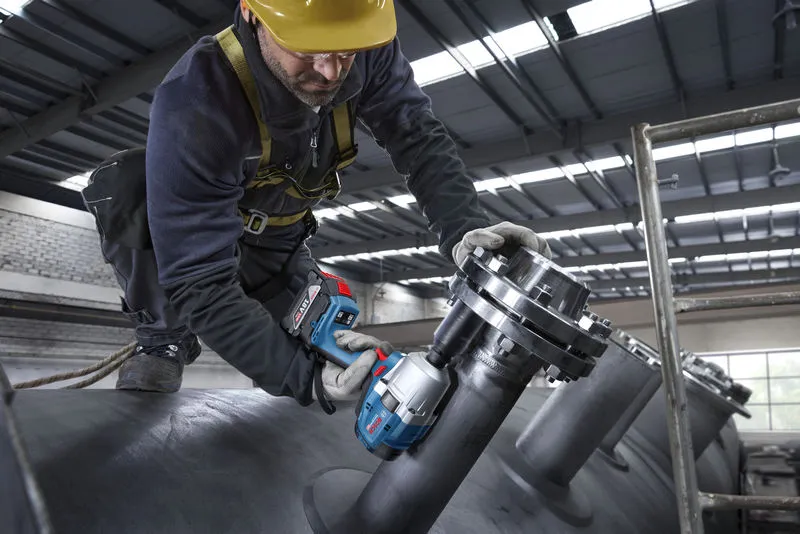 Una persona che indossa dispositivi di sicurezza utilizza una chiave a impulsi a batteria su tubazioni industriali.