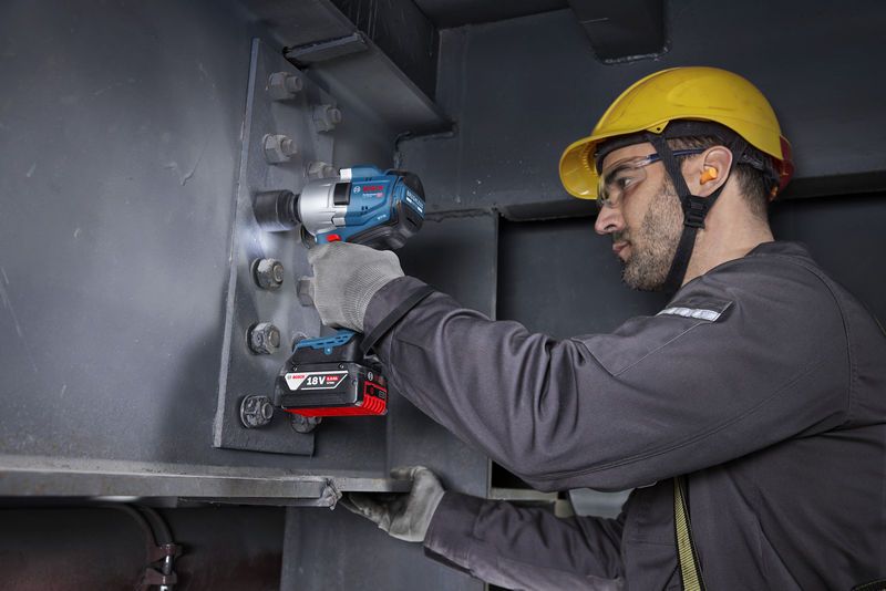 Una persona che indossa dispositivi di sicurezza stringe i bulloni su una trave di acciaio utilizzando una chiave a impulsi a batteria.