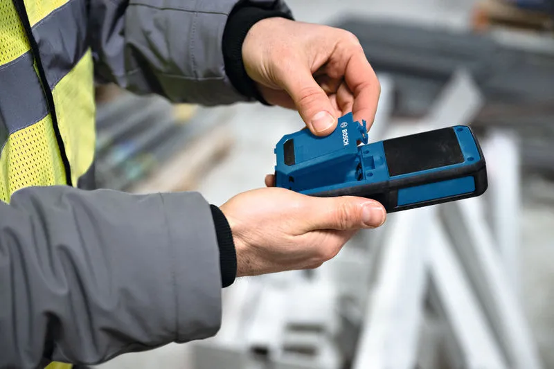 Una persona che indossa un equipaggiamento di sicurezza apre il vano batteria di un misuratore di umidità dei materiali.