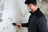 Una persona che indossa un equipaggiamento di sicurezza utilizza un misuratore di umidità del materiale su una parete danneggiata.