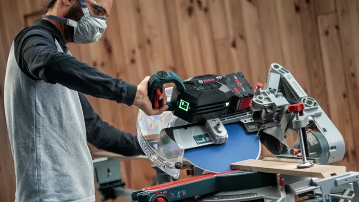 Una persona che indossa dispositivi di sicurezza aziona una sega troncatrice per tagliare una tavola di legno.