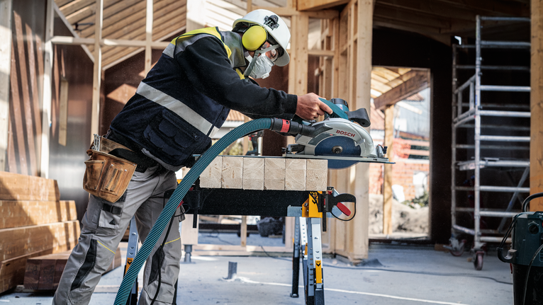 Una persona che indossa dispositivi di sicurezza taglia travi di legno con una sega circolare in un cantiere edile.