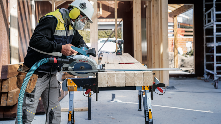 Una persona che indossa dispositivi di sicurezza taglia travi di legno con una sega circolare in un cantiere edile.